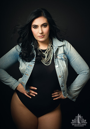 Contemporary studio portrait of a young woman in a black bodysuit, denim jacket and silver necklaces looking at the camera by Vancouver portrait photographer Angela McConnell