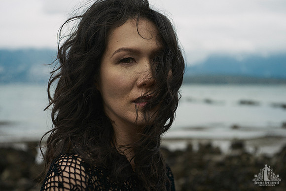 Environmental portrait of an asian woman standing in front of a rocky shore looking at the camera with her hair being blown across her face by Vancouver portrait photographer Angela McConnell