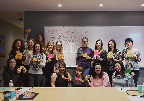 A group of women pose together in front of a white board with empowering phrases, each holding a small neon card with a phrase on them