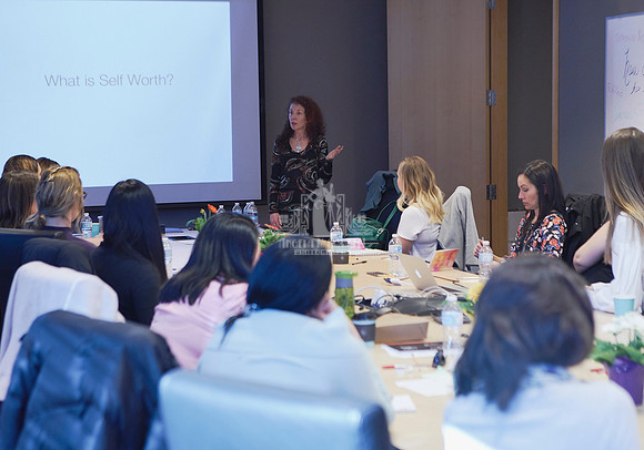 A presenter stands at the head of a board room table surrounded by women listening to her presentation during a workshop